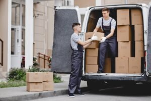 Two young handsome movers wearing uniforms are unloading the van full of boxes. House move, mover service.